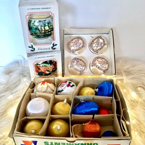 May include: A box of 12 vintage hand-decorated Christmas tree ornaments. The ornaments are in various colors and designs, including gold, red, blue, and white. The box is labeled "Hand decorated Christmas tree ornaments".
