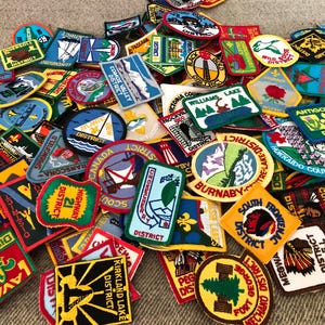 May include: A collection of colorful embroidered patches, featuring various shapes and designs. Many patches display text such as "District", "Canada", and "Williams Lake". The patches are in various colors, including red, blue, green, and yellow.