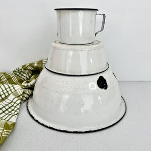 May include: A stack of three white enamelware bowls and a mug with black trim. The bowls decrease in size, with the largest at the bottom. A green and white checkered cloth is visible on the left.