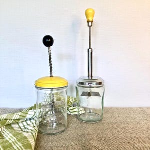 以下が含まれることがあります： 2つのヴィンテージガラスフードチョッパー。1つは黄色の蓋と黒いノブ、もう1つはシルバーの蓋と木製のノブが付いています。どちらにもガラスに測定マークが付いています。緑と白のチェック柄の布が背景にあります。