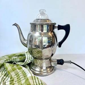 May include: A polished silver-tone electric percolator coffee pot with a clear glass knob on the lid, a curved spout, and a black handle. The base features a black electrical cord attachment extending to the right side of the metallic body.
