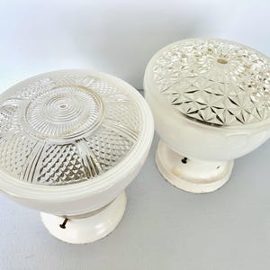 May include: Two vintage white and clear glass light fixtures. Each fixture has a white base and a clear glass dome. One dome has a spiral design, and the other has a diamond pattern. The fixtures are likely for ceiling or wall mounting.