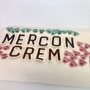 May include: A collection of green and red felt patches with numbers on them. The patches are arranged in a semi-circle around the word "MERCON CREM" in black and gold felt letters.