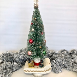 May include: A vintage Christmas tree made of green bottle brush with a white star on top, decorated with colourful ornaments and a red bow. The tree is sitting in a white sleigh with gold trim and is surrounded by silver tinsel.