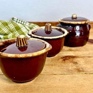 May include: Three brown ceramic bowls with lids, featuring a glossy finish and a contrasting tan rim. The lids have a textured, cone-shaped handle. The bowls are arranged on a wooden surface, with a green and white checkered cloth in the background.