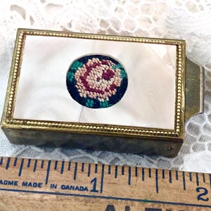 May include: An antique brass box with a mother-of-pearl top. The top has a circular, embroidered floral design in pink, green, and blue. The box is approximately 5 cm long and has a hinged closure.