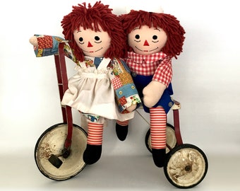 May include: Two red-haired rag dolls riding a red and white tricycle. The dolls are wearing colorful patchwork dresses and have red and white striped stockings.