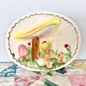 May include: A ceramic wall plaque featuring a girl with blonde hair and a red bow sitting under a large, yellow mushroom with a pink mushroom in the foreground. The plaque has a white background with green grass and red flowers.