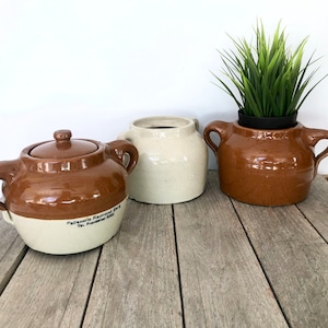 May include: Three brown and white ceramic crocks with two handles each. The crock on the left has a lid and the text "Patisserie Raymond Berg Tel. Providence 5161" on the side. The crock on the right has a green plant inside.