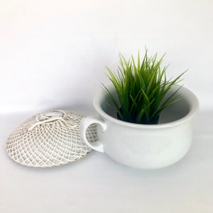 May include: White ceramic chamber pot with a white crocheted lid and a green plant inside.