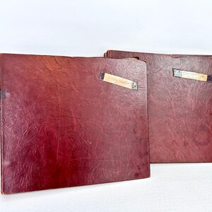 May include: Two vintage burgundy-colored leather-bound books. The books have a textured surface and are rectangular in shape. Each book has a rectangular label adhered to the spine. The books are placed on a white surface.
