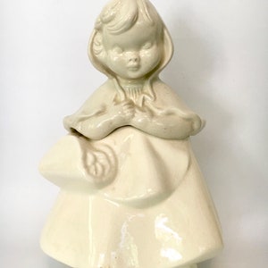 May include: White ceramic cookie jar shaped like a girl wearing a red hooded cloak. The girl is holding a basket in her left hand.