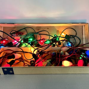 May include: A string of multi-coloured incandescent Christmas lights with dark green wiring, coiled inside an open rectangular cardboard box. The bulbs are red, green, blue, orange, and white, featuring a classic smooth, teardrop-shaped glass design.