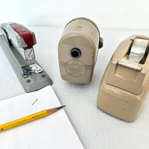 May include: An assortment of vintage office supplies, including a gray and red stapler, a tan pencil sharpener with a star logo, and a tan tape dispenser. A yellow pencil and a piece of paper are also present.