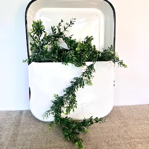 Può includere: Fioriera da parete in smalto bianco con bordo nero, riempita di finto fogliame verde. La fioriera ha una forma rettangolare con fondo curvo e manico. L'estetica generale è in stile rustico.