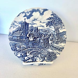 Può includere: Un piatto decorativo blu e bianco con una scena rurale. Il piatto presenta una carrozza trainata da cavalli che attraversa un ponte, una chiesa e degli alberi. Il piatto è rotondo con un bordo smerlato.