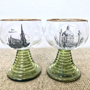 May include: Two clear glass wine glasses with gold rims and green stems. Each glass features a black and white illustration of a famous building. One glass shows the Stephansdom in Vienna, Austria. The other glass shows the dome of St. Paul's Cathedral in London, England.