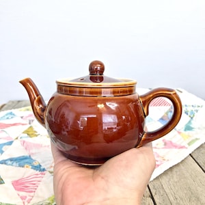 May include: A small brown ceramic teapot with a rounded body and a single handle. The teapot has a lid with a small knob on top.