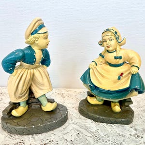 Peut inclure: Deux figurines en céramique représentant des enfants hollandais en costume traditionnel. Le garçon porte une veste bleu sarcelle, un pantalon beige et des sabots jaunes. La fille a une robe bleu sarcelle et jaune. Les deux sont sur des socles gris.