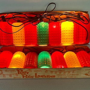 May include: A set of Party Patio Lanterns in a cardboard box, featuring two rows of cylindrical, textured plastic light covers in red, yellow, and green. The black electrical cord connects the illuminated lanterns, which are arranged in a display.