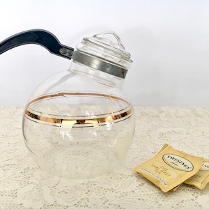 May include: A clear glass teapot with a black plastic handle and gold trim. The teapot is sitting on a white lace tablecloth. There are three tea bags in the image, labeled "Twinings Earl Grey Tea".