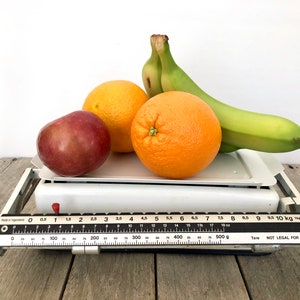 May include: A vintage white scale with a black and white measurement bar, displaying a red apple, two oranges, and a bunch of green bananas. The scale reads up to 10 kg / 22 lbs.
