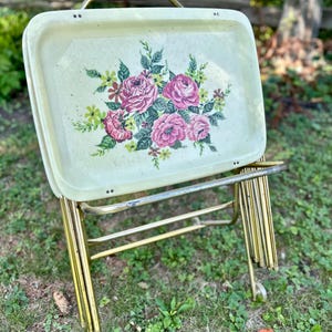 May include: Vintage serving tray with a floral design featuring pink roses and green foliage. The tray is set on a gold-colored metal stand with wheels, and a wooden handle. A charming piece for serving or display.