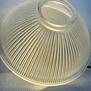 May include: A clear glass lampshade with a ribbed design. The lampshade has a rounded, bowl-like shape with a circular opening at the top. The ribbed pattern creates a textured surface, and the glass appears to be slightly frosted.
