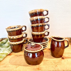 May include: A vintage brown ceramic coffee set, including cups, saucers, a creamer jug, and a lidded sugar bowl. The cups and sugar bowl feature a two-tone glaze with a lighter brown rim. The items are arranged on a wooden surface.