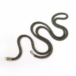 Peut inclure: Un collier de perles vert foncé avec un design en forme de serpent. Le collier est fait de petites perles rondes et a un fermoir argenté.