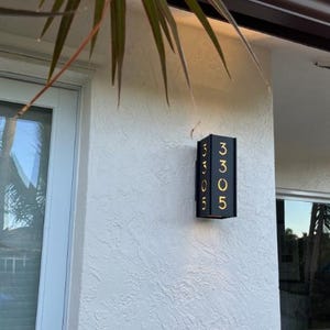 May include: A black metal house number plaque with gold numbers, displaying the number 3305. The plaque is mounted on a white stucco wall.