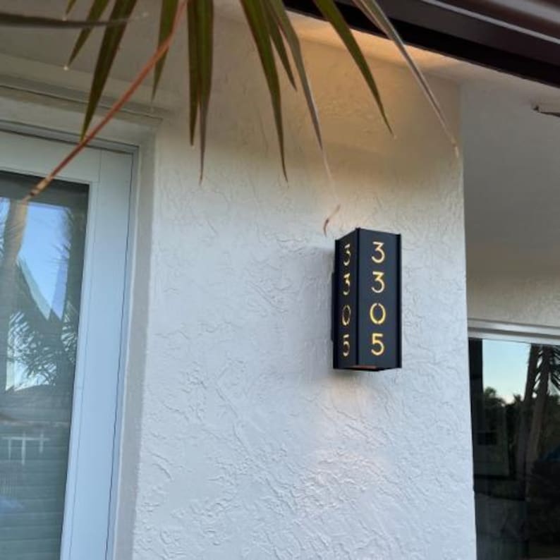 May include: A black metal house number plaque with gold numbers, displaying the number 3305. The plaque is mounted on a white stucco wall.