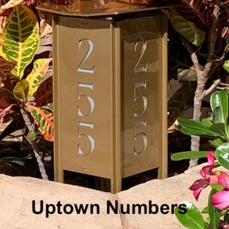 May include: A gold metal house number plaque with the number 255 displayed on both sides. The plaque is mounted on a metal stand with a square base. The text "Uptown Numbers" is visible below the plaque.