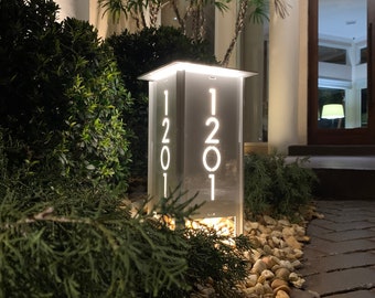 Könnte beinhalten: Ein modernes, beleuchtetes Hausnummernschild mit der Nummer 1201 in Weiß auf grauem Hintergrund. Das Schild ist von innen beleuchtet und von einem Bett aus kleinen Steinen umgeben.