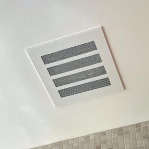 May include: A square, white ceiling vent with four horizontal, rectangular black mesh openings. The vent is set into a white ceiling, with a glimpse of a tiled wall in the lower portion of the image.