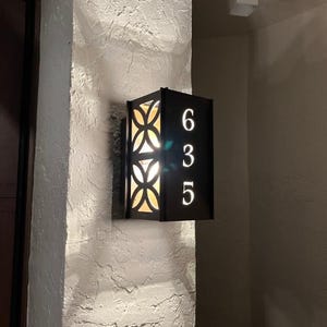 May include: Black metal house number plaque with a geometric design and the numbers 635. The plaque is illuminated from behind, casting a warm glow.