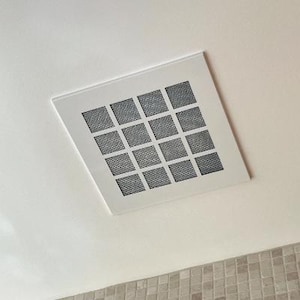 May include: A square, white ceiling vent with a grid of sixteen black, square mesh openings. The vent is set into a white ceiling, with a glimpse of a tiled wall at the bottom.