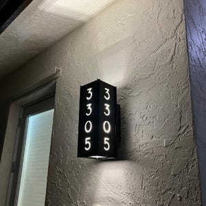 May include: Black metal house number plaque with white illuminated numbers 3305. The plaque is mounted on a white wall.
