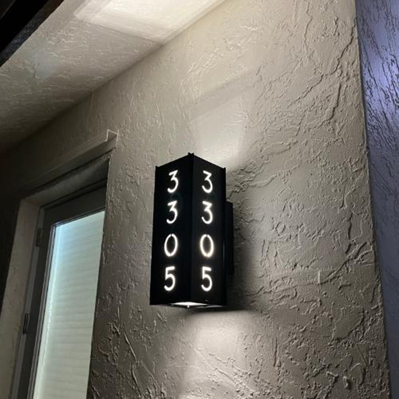 May include: Black metal house number plaque with white illuminated numbers 3305. The plaque is mounted on a white wall.