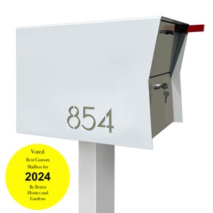 May include: White metal mailbox with a red flag and a silver door. The mailbox has a white post and the number 854 is cut out of the front. A yellow circle sticker on the post reads "Voted Best Custom Mailbox for 2024 By Better Homes and Gardens".