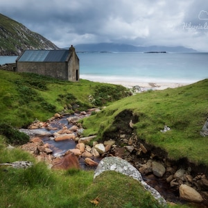 May include: A small stone building sits on a grassy hillside overlooking a beach with a clear blue ocean. A small stream flows down the hillside in front of the building.