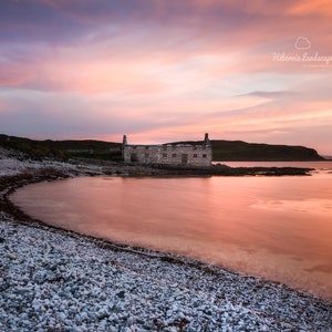 Può includere: Fotografia di un paesaggio costiero al tramonto. L'immagine presenta un edificio in pietra su una piccola isola, una spiaggia di ciottoli e acqua calma che riflette le tonalità rosa e arancioni del cielo nuvoloso.