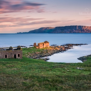 Può includere: Una vista panoramica di una costa rocciosa con due rovine di pietra in primo piano. Le rovine sono parzialmente crollate e ricoperte di vegetazione. Il cielo è di un rosa e un blu delicato, e l'acqua è di un blu calmo. In lontananza, una grande scogliera si erge dall'acqua.