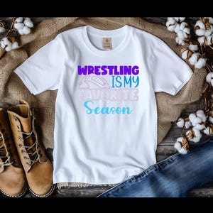 May include: White t-shirt with the text "Wrestling is my Favorite Season" in purple, pink, and blue. The shirt is displayed on a rustic wooden surface with brown boots, denim jeans, and cotton sprigs.
