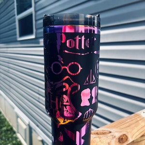 Puede incluir: Vaso negro con un degradado de rosa, morado y naranja. El vaso presenta la palabra "Potter" y otros gráficos. El vaso está sobre una superficie de madera.