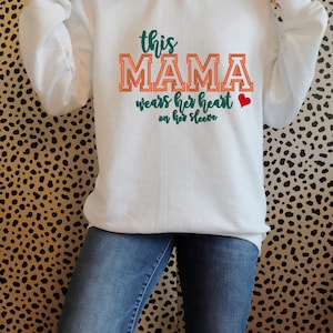 May include: White crewneck sweatshirt with the text "This Mama wears her heart on her sleeve" in green and orange lettering, with a small red heart. The sweatshirt is paired with blue jeans.