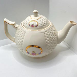 May include: Cream-colored ceramic teapot with a diamond pattern and gold trim. The teapot features a Claddagh design with a heart, crown, and hands. The handle and spout are curved, and the lid has a small finial.