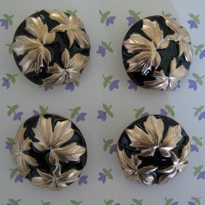 Könnte beinhalten: Vier runde, schwarze Knöpfe mit goldenen Blattmotiven. Die Knöpfe sind rund und haben ein erhabenes Design.