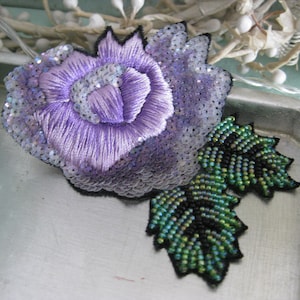 Könnte beinhalten: Eine violette und grüne Perlenblumenbrosche. Die Blume besteht aus Pailletten und Stickerei. Die Blätter sind aus grünen Perlen.