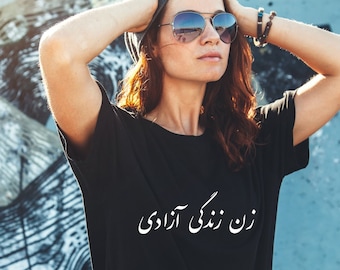 以下が含まれることがあります： 黒いTシャツを着た女性。Tシャツにはペルシャ語で「زن زندگی آزادی」（女性、人生、自由）と白文字で書かれています。彼女はサングラスをかけており、両手で頭を支えています。